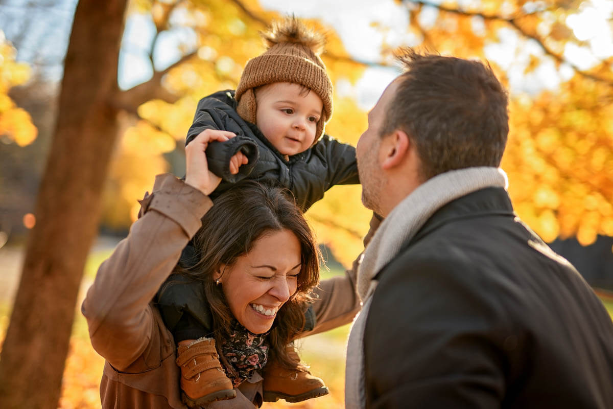 A family of three having fun in a park. One adult has a toddler on their shoulders and is laughing facing the other adult.