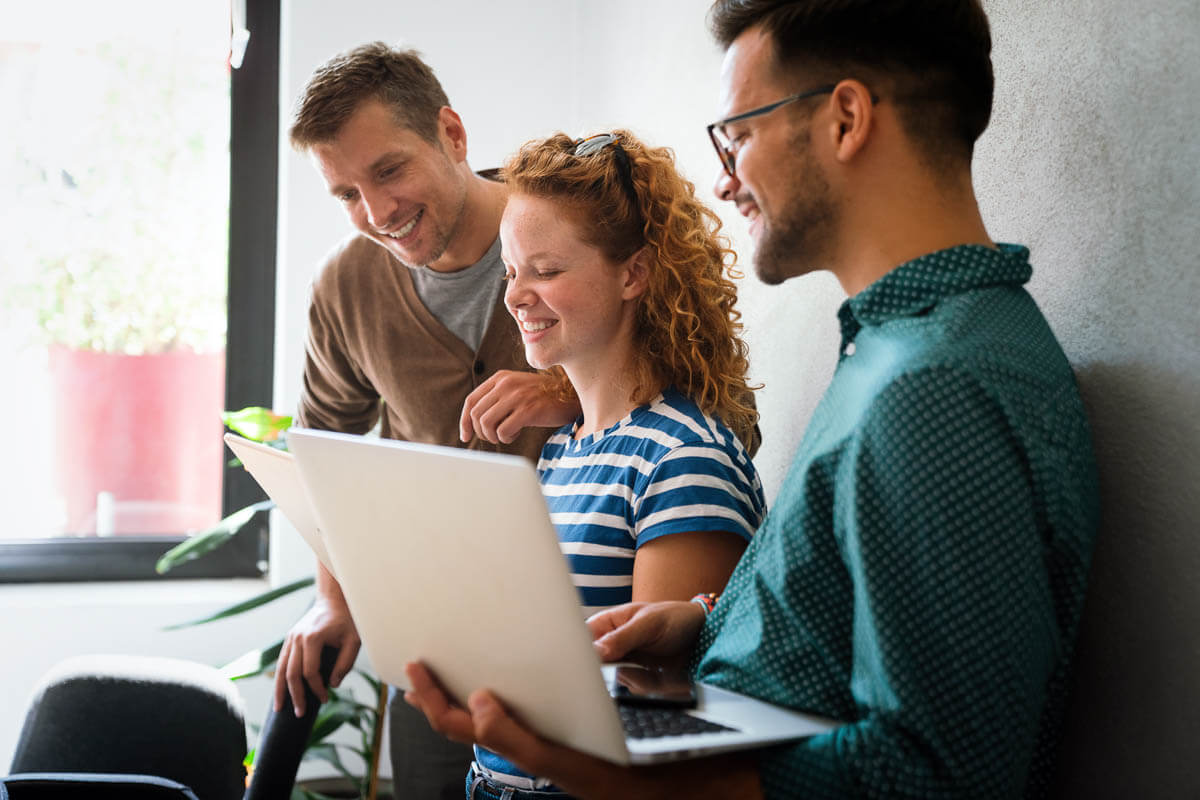 Three smiling colleagues leaning against a wall looking at a laptop