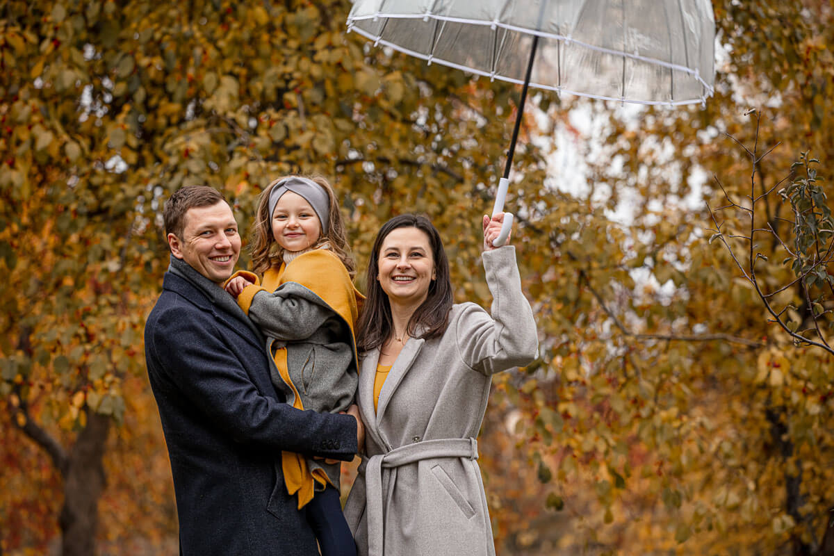 Family of three in front of autumnal trees. One adult has the child in his arms while the other holds an umbrella above them.