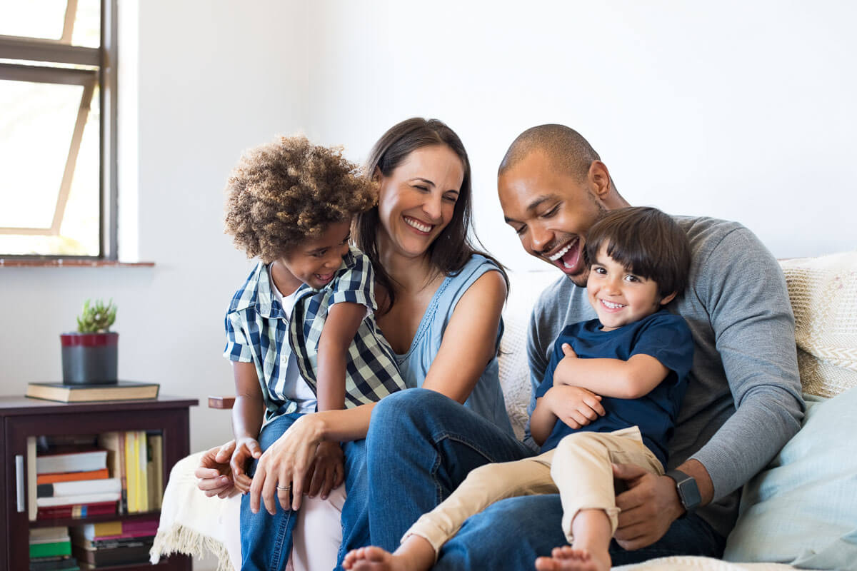 A diverse family with two young children sitting on their sofa, each adult holding a child on their lap.
