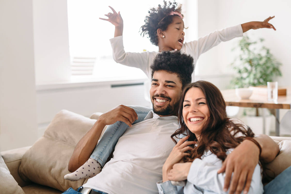 A family of three sitting on their sofa. The child is sitting on the back of the sofa above one of the adults while striking a pose.