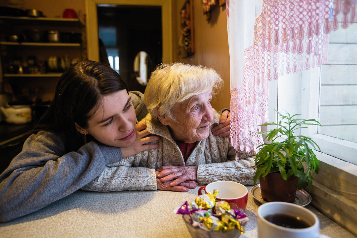 An elderly adult is sat at their kitchen table looking out of the window while their grand child is hugging their shoulders from behind.