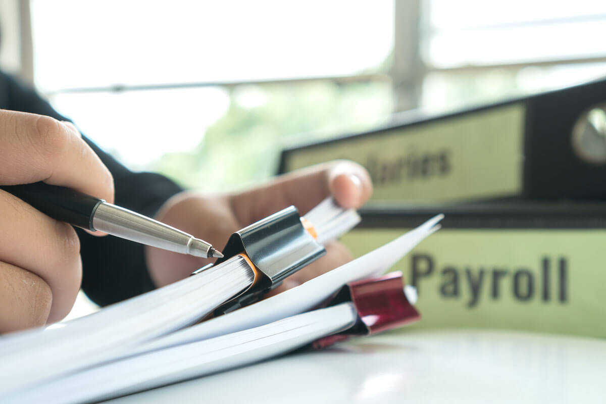 Person at a desk with two clipped documents next to two box files, one of which is marked 'Payroll'