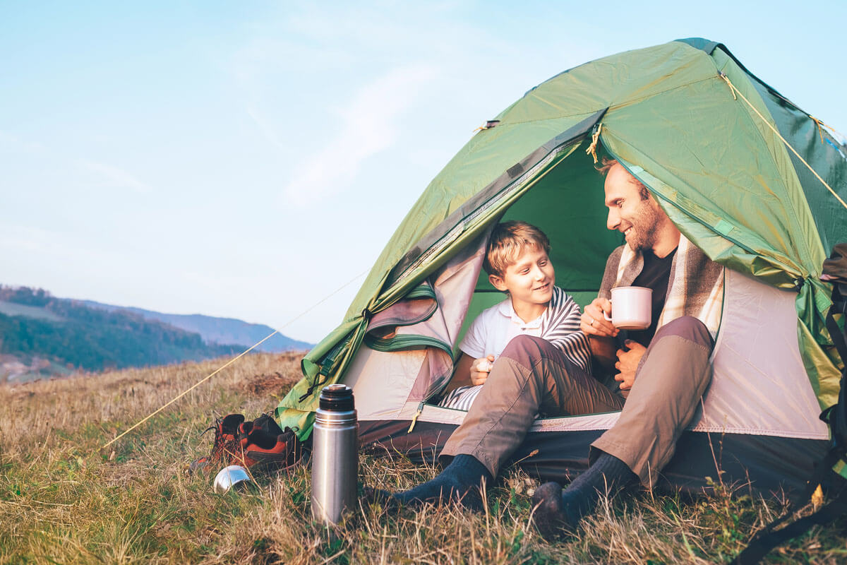 A parent and child smiling at each other while they sit in the doorway of their tent in the hills. The parent is holding a mug.