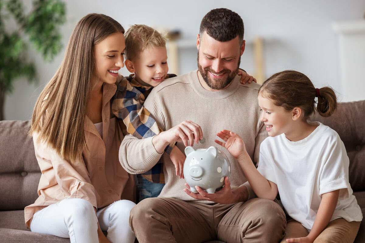 Family of four sat on a sofa. One adult is putting coins into a white piggy bank, helped by the older child, while the other adult and younger child watch on.
