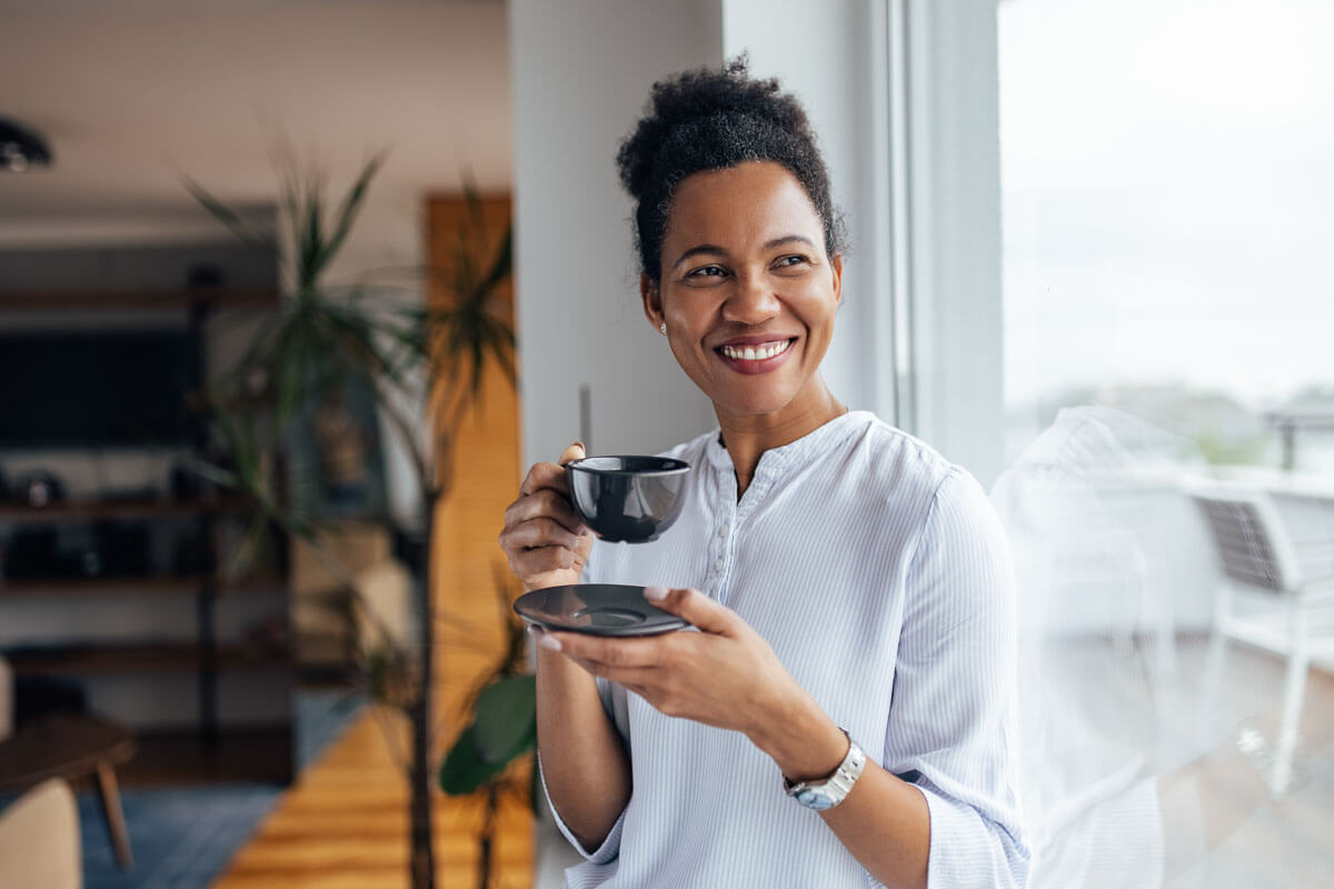 Smiling woman next to a window, holding a cup in her right hand and saucer in her left.