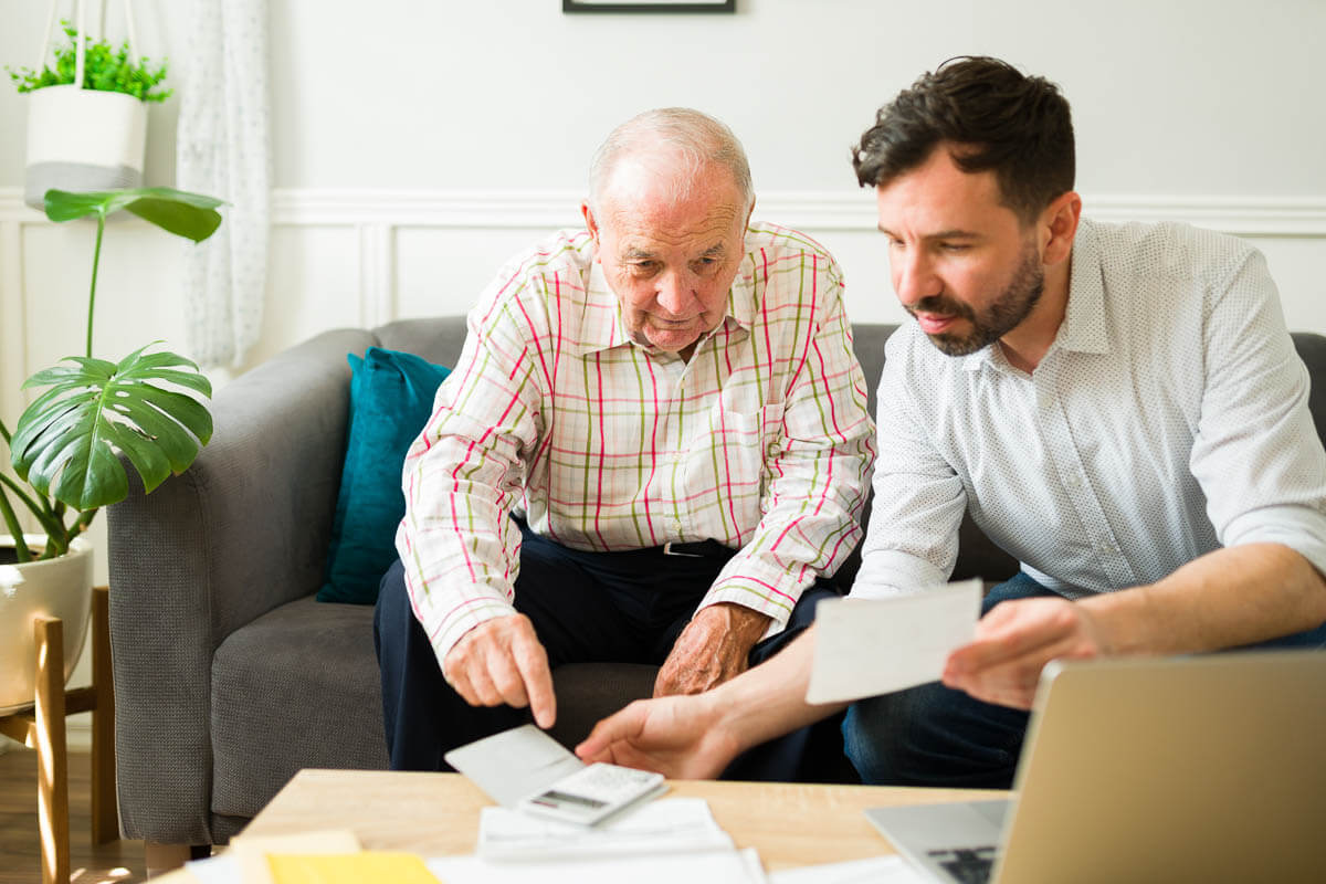 Elderly adult on the sofa with a younger adult looking at papers, a calculator and laptop on the coffee table.