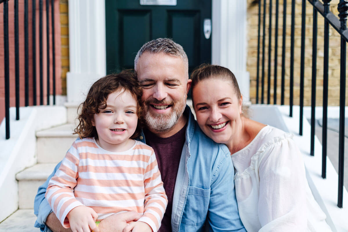 Family of three sitting on their front steps, with a child is on one adult's knee and the other adult sitting behind them.