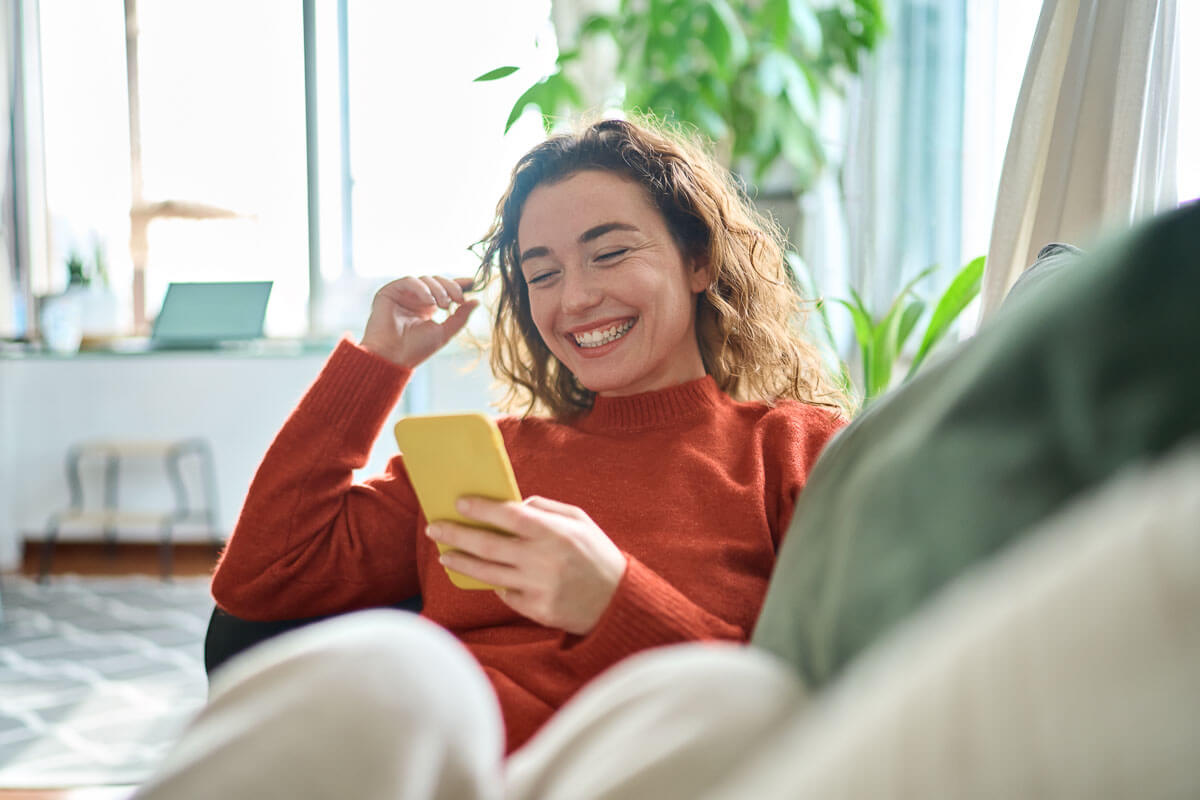 A young woman is lying on her sofa looking at her mobile phone and smiling broadly.