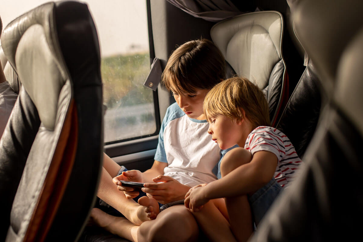 Two siblings on a coach playing a video game on a mobile phone. The older child is cross-legged and holding the phone while the younger child watches on.