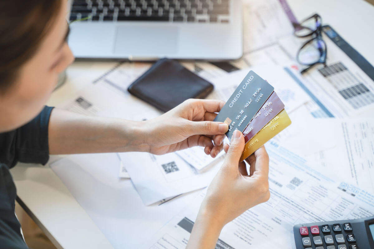 A woman is sat at a desk surrounded by bills, paperwork, a calculator, laptop and her glasses. She is holding three credit cards.